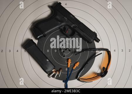 A shooting target with bullet holes in the center, a G19gen5 pistol, goggles and earplugs, and a magazine with cartridges. Stock Photo
