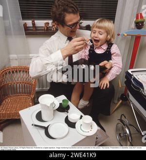 In the 1960s. A father playing with his daughter, having a chocolate ball on a spoon, ready to go inte her mouth. In front of them a toy kitchen range with cups and saucers on top. A playroom or childrens room with a pram and a childrens sized chair. 1969. Kristoffersson Stock Photo