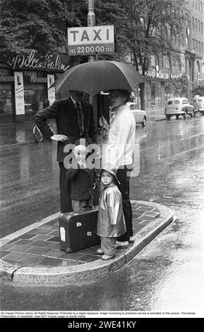 Stockholm in the 1950s. It is a rainy day in central Stockholm. A young couple stands under an umbrella at a taxi pole and waits for a taxi but it can be a long wait under the umbrella for the young couple in the rainy weather. Only one of the children is wearing rain gear. The boy is caught in the picture like many other children, picking his nose. Stockholm 1953. Kristoffersson ref 2A-12 cv26-3 Stock Photo