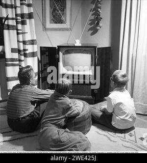 Television set in the 1950s. An early television set in period design wooden cabinett with only the ability to display television broadcasts in black and white. The antenna is seen standing on the tv.  The knobs on the front control the picture and sound. The three boys in front of it looks at it in fascination. 1956 Svahn ref SVA1 Stock Photo