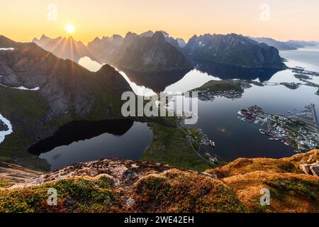 Sunset casts radiant light over Reinebringen, with the sun mirroring on calm fjord waters, the moss-covered foreground leading to Reine Stock Photo