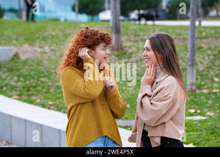 Close-up of two joyful women engaging in a lively conversation, with one touching her glasses, in a green urban park Stock Photo