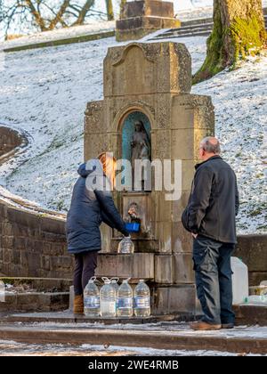 Woman filling plastic bottles with water from St Ann's well at the foot of The Slopes, in winter, in snow. Buxton, UK Stock Photo