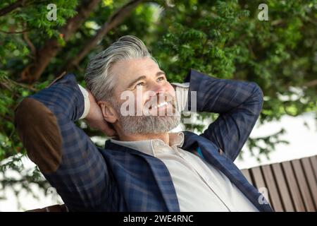 A 45 year old caucasian male in a dark blue blazer and white t-shirt, relaxing outdoors with a contented smile, exuding a serene and happy mood. Stock Photo