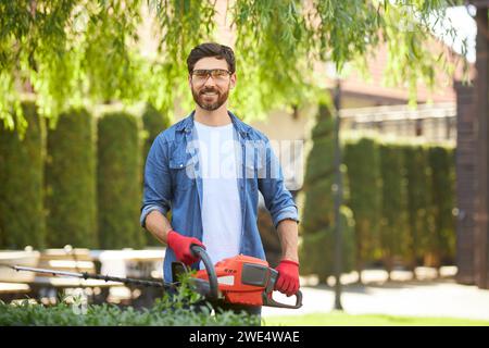 Smiling handyman in casual clothes looking at camera, while working in garden. Portrait of bearded worker using electric hedge trimmer, taking care of evergreen plants. Concept of seasonal work. Stock Photo