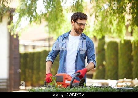 Smiling brunet male gardener cutting overgrown bush by hedge trimmer at sunny day. Front view of hard working man using modern gardening equipment for seasonal work, trimming hedges. Concept of work. Stock Photo