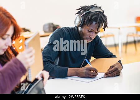 Young African male student with headphones intently writing notes at a classroom desk, absorbed in study. Stock Photo