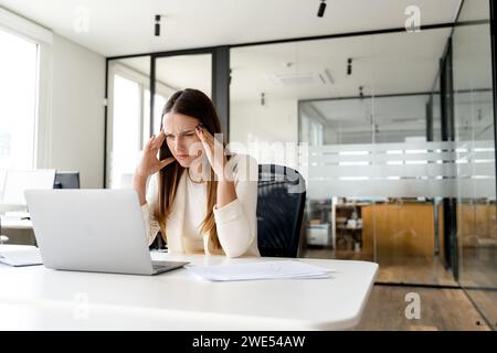 Overwhelmed by the demands of her work, a businesswoman in a light suit presses her temples, trying to concentrate amidst the stress. The taxing moments of corporate life Stock Photo