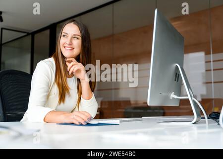 A young businesswoman is captured seated at her desk, exuding confidence and contentment as she interacts with a computer in sleek, contemporary office. Smiling female office employee enjoying her job Stock Photo