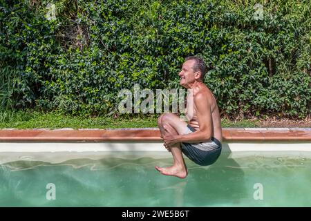 Caucasian man with a funny expression takes a bomb dive in the swimming pool on a sunny day Stock Photo