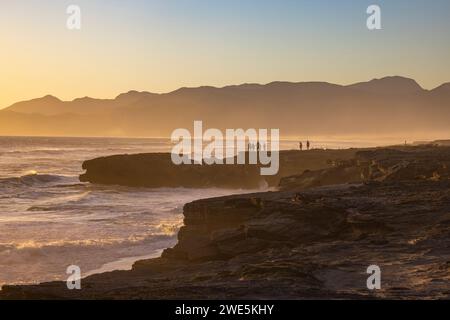 People with fat tire bicycles stand on rocks as waves break onto the coast and beach at sunset in Walker Bay Nature Reserve, Gansbaai De Kelders, West Stock Photo