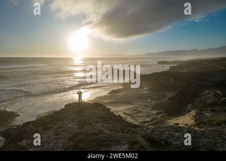 Aerial view and silhouette of a man standing on a rock overlooking the coast and beach at sunset in Walker Bay Nature Reserve, Gansbaai De Kelders, We Stock Photo