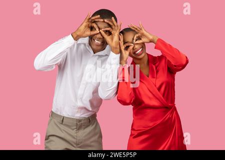 Couple making fun hand gestures over eyes, playful poses, pink background Stock Photo