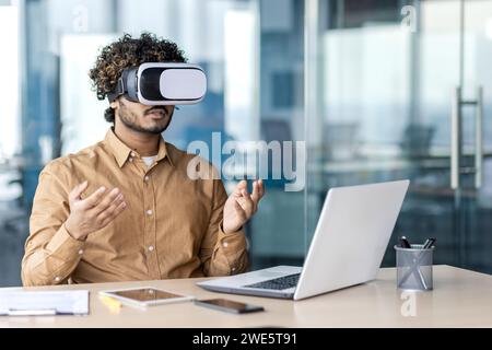 Indian man in VR glasses wearing sandy shirt while sitting by desktop with open laptop and raising hands in office. Creative digital designer using 3d innovation for working in cyber database. Stock Photo