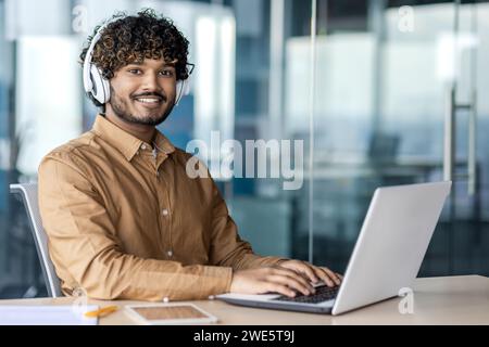 Indian guy with curly hair sitting in wireless headphones at working place with laptop and looking at camera. Confident man listening to playlist for improving focusing on project enhancement indoors. Stock Photo