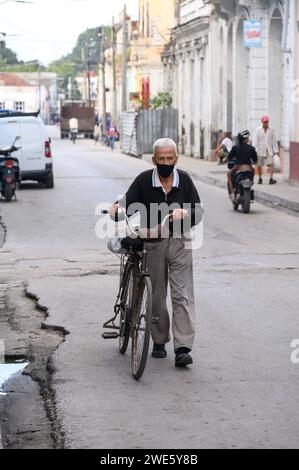 A senior man with face mask pushes a bicycle by black water in the street curb. Stock Photo
