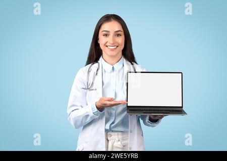 Happy woman doctor in white lab coat presenting laptop with blank screen, suitable for medical software interface Stock Photo