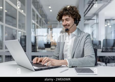Smiling young businessman with headset working on laptop in modern office, showcasing multitasking and customer service skills. Positive work environment reflected in his cheerful expression. Stock Photo