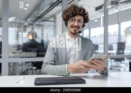 Cheerful young businessman in a headset at work, displaying professionalism, competence, and a positive emotional state in a bright office environment. Stock Photo