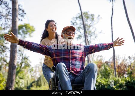 Social worker pushing cheerful retired senior man with arms outstretched in wheelchair at park Stock Photo