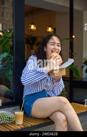 Smiling woman sitting and eating burger on bench Stock Photo