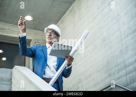 Engineer examining and holding tablet PC with blueprint near wall Stock Photo