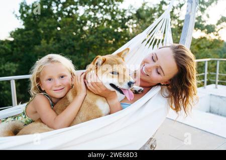 Smiling mother and daughter petting Shiba Inu dog in hammock Stock Photo