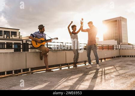 Happy man playing guitar with friends dancing on rooftop Stock Photo