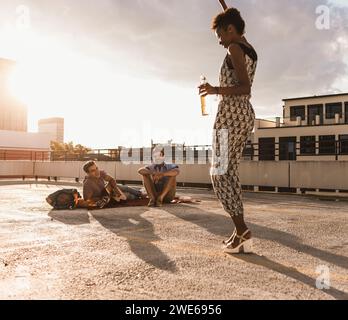 Happy woman holding beer bottle and dancing in front of friends on rooftop Stock Photo