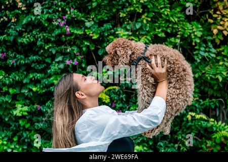 Happy young woman playing with pet poodle dog in front of plants Stock Photo