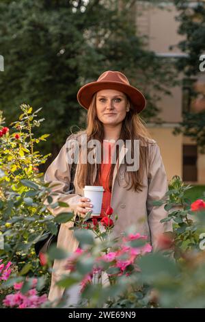 Smiling young woman wearing hat and standing with coffee cup near flowering plants Stock Photo