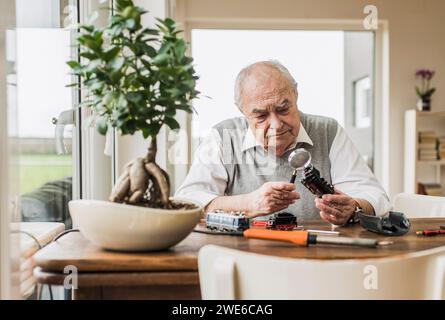 Senior man sitting near window and examining miniature train through magnifying glass at home Stock Photo