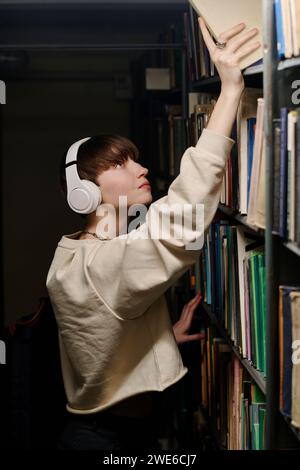 Young student wearing wireless headphones and taking book from top shelf in library Stock Photo