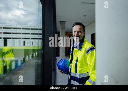 Smiling engineer holding hardhat and standing near glass window Stock Photo