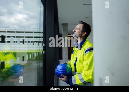 Contemplative engineer standing near window with hardhat in office Stock Photo