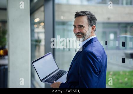 Happy businessman holding laptop near glass window in office Stock Photo