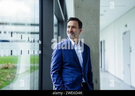 Smiling mature businessman looking through window and standing in office corridor Stock Photo