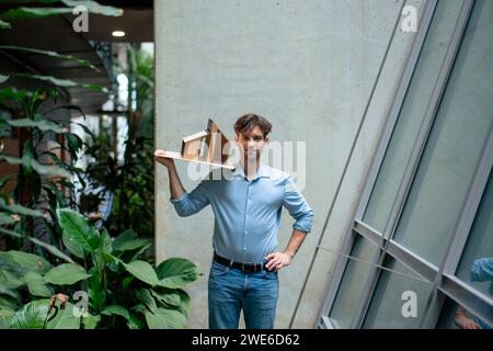 Young architect with hand on hip holding model house in office Stock Photo
