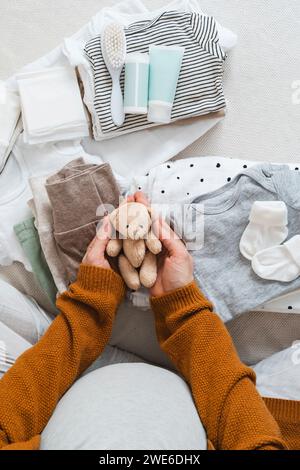 Pregnant woman holding stuffed teddy bear near clothes at home Stock Photo