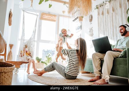 Mother playing with her son in living room at home with father using laptop Stock Photo