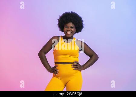 Happy young woman in yellow sports clothing standing with arms akimbo against two tone colored background Stock Photo