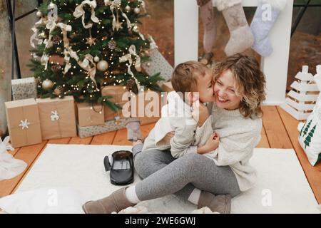 Boy kissing smiling mother sitting on carpet near Christmas tree at home Stock Photo