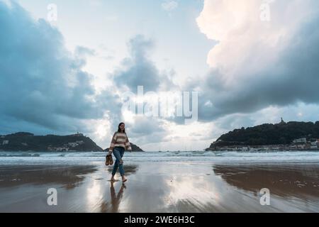 Young tourist walking on Playa de la Concha shore at sunset under cloudy sky Stock Photo
