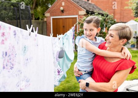 Happy woman carrying daughter near clothes hanging on clothesline in backyard Stock Photo