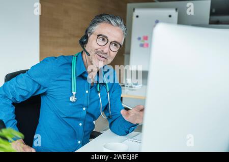 Mature doctor wearing headset and talking on video call through computer Stock Photo