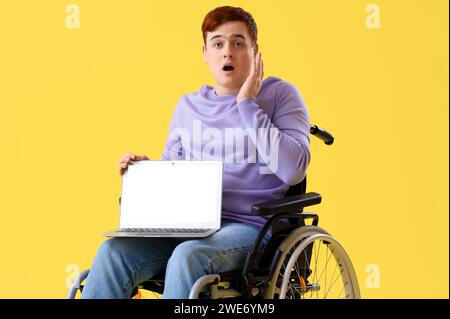 Shocked young man in wheelchair with laptop on yellow background Stock Photo