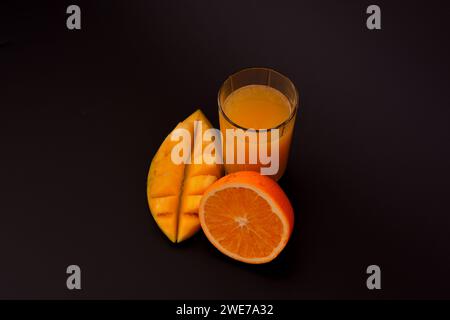 A mixture of tropical fruit juice in a tall faceted glass on a black background, next to it are ripe mango halves and an orange. Close-up. Stock Photo