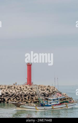 Small fishing trawler passing in front of red lighthouse as it enters harbor in South Korea Stock Photo