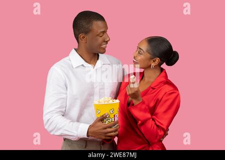 Black man and woman sharing popcorn, enjoying moment, dressed in white and red elegant attire Stock Photo
