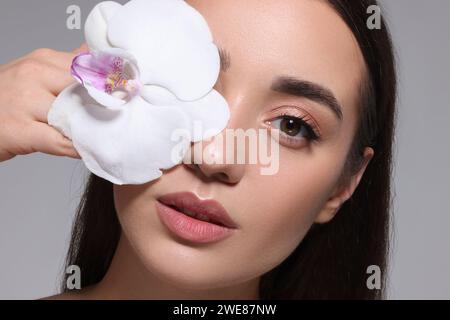 Beautiful young woman covering eye with orchid flower on grey background, closeup Stock Photo